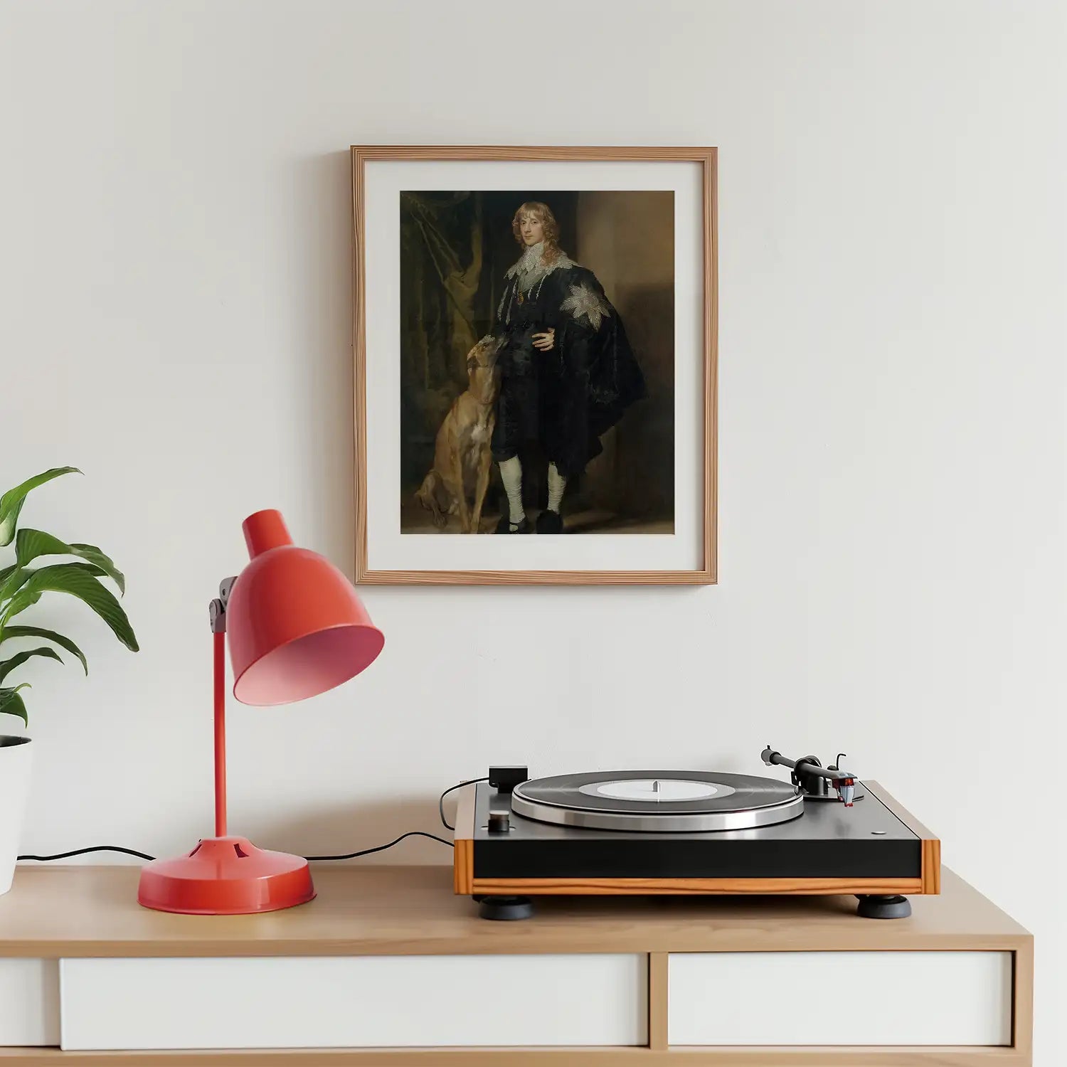 A framed painting of a man in historical clothing standing next to a dog, displayed on a wall above a record player and a red desk lamp, with a green plant nearby.