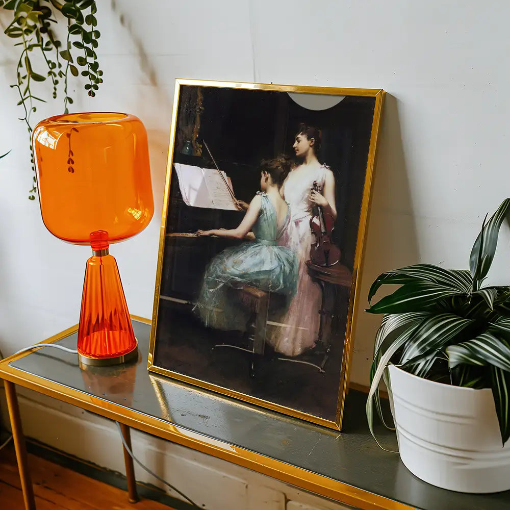Framed painting of two women playing music, one at a piano and the other holding a violin, displayed on a table with an orange lamp and a potted plant.