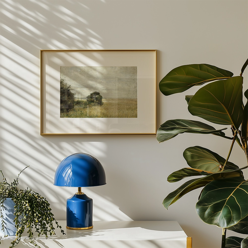 Framed landscape painting above a blue lamp and green plant on a white table against a wall with sunlight shadows.