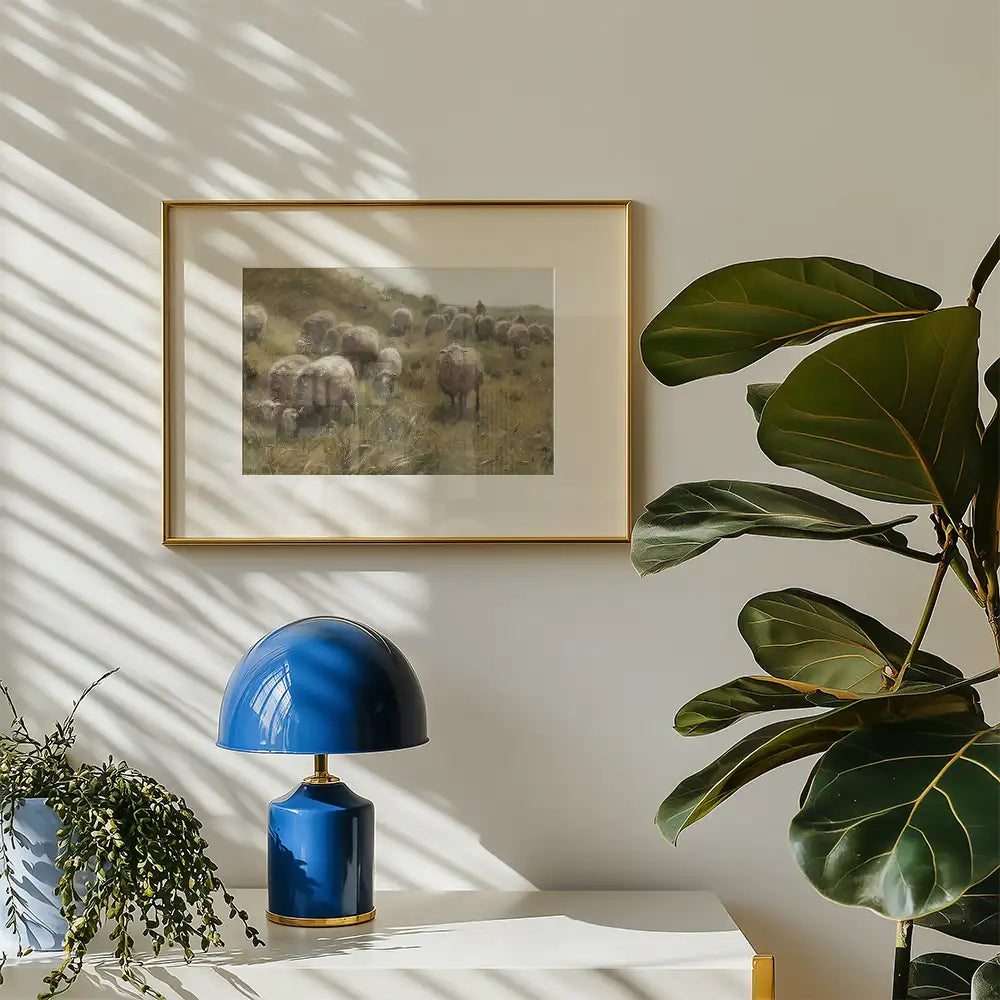 Framed painting of a herd of sheep in a natural setting, displayed on a light wall with a decorative blue lamp and lush green plant nearby.
