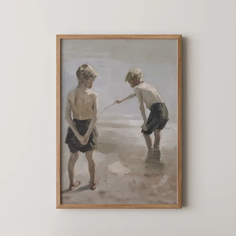 Painting of two boys playing at the beach, one standing and looking on while the other crouches and interacts with the water, framed in a light wood frame.