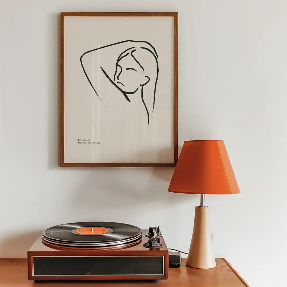 Modern line art poster of a woman with long hair, framed and displayed beside a vintage record player and an orange lampshade, perfect for minimalist home decor.
