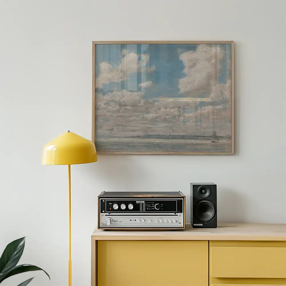 Framed painting of a cloudy sky above a serene ocean, displayed above a yellow lamp and a vintage audio system on a wooden cabinet.