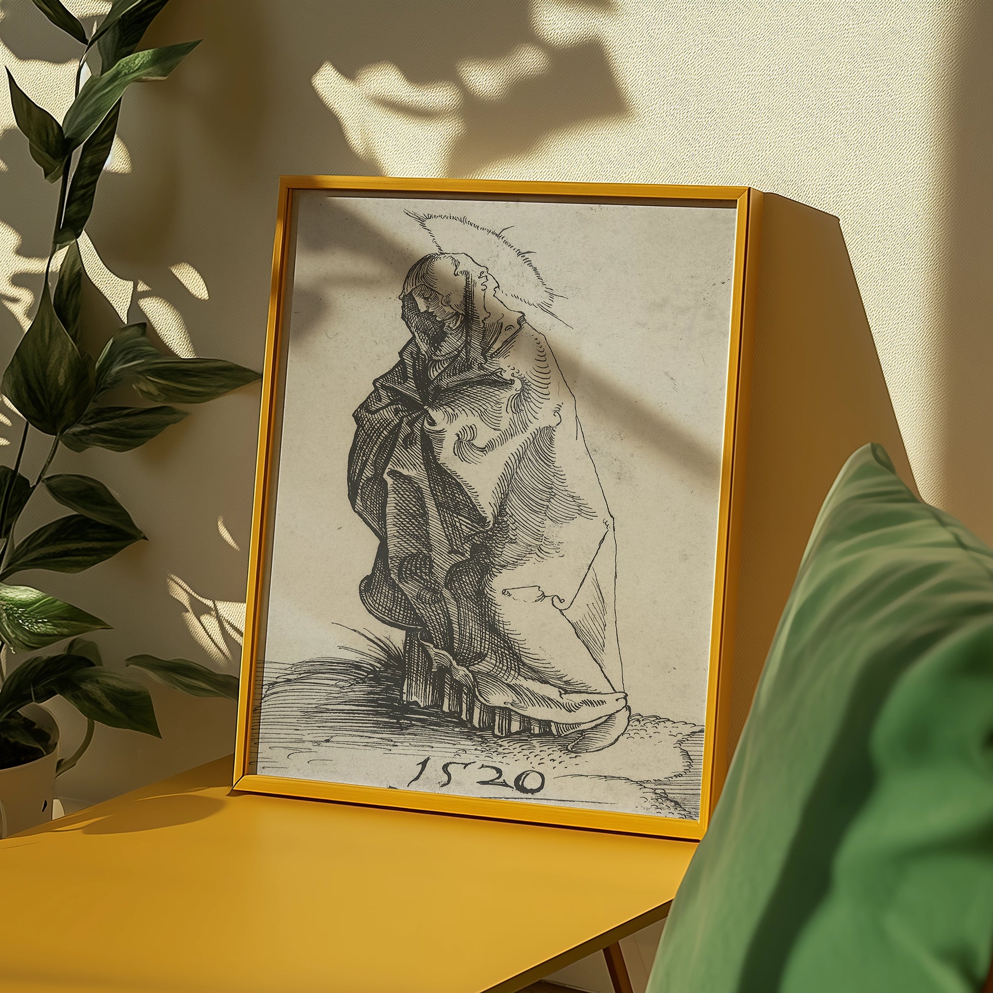 Framed vintage drawing of a cloaked figure with the year 1520, displayed on a yellow table beside green cushions and foliage.