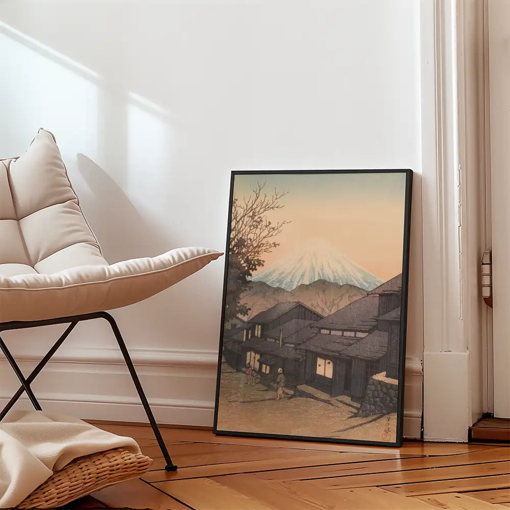 Framed painting of a serene landscape featuring a mountain and traditional houses, leaning against a wall beside a modern chair.