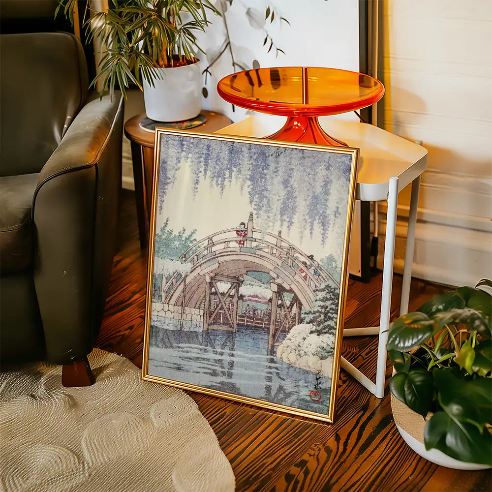 Framed painting of a serene landscape featuring a bridge over water, with greenery and snow, placed beside a chair and a small table.