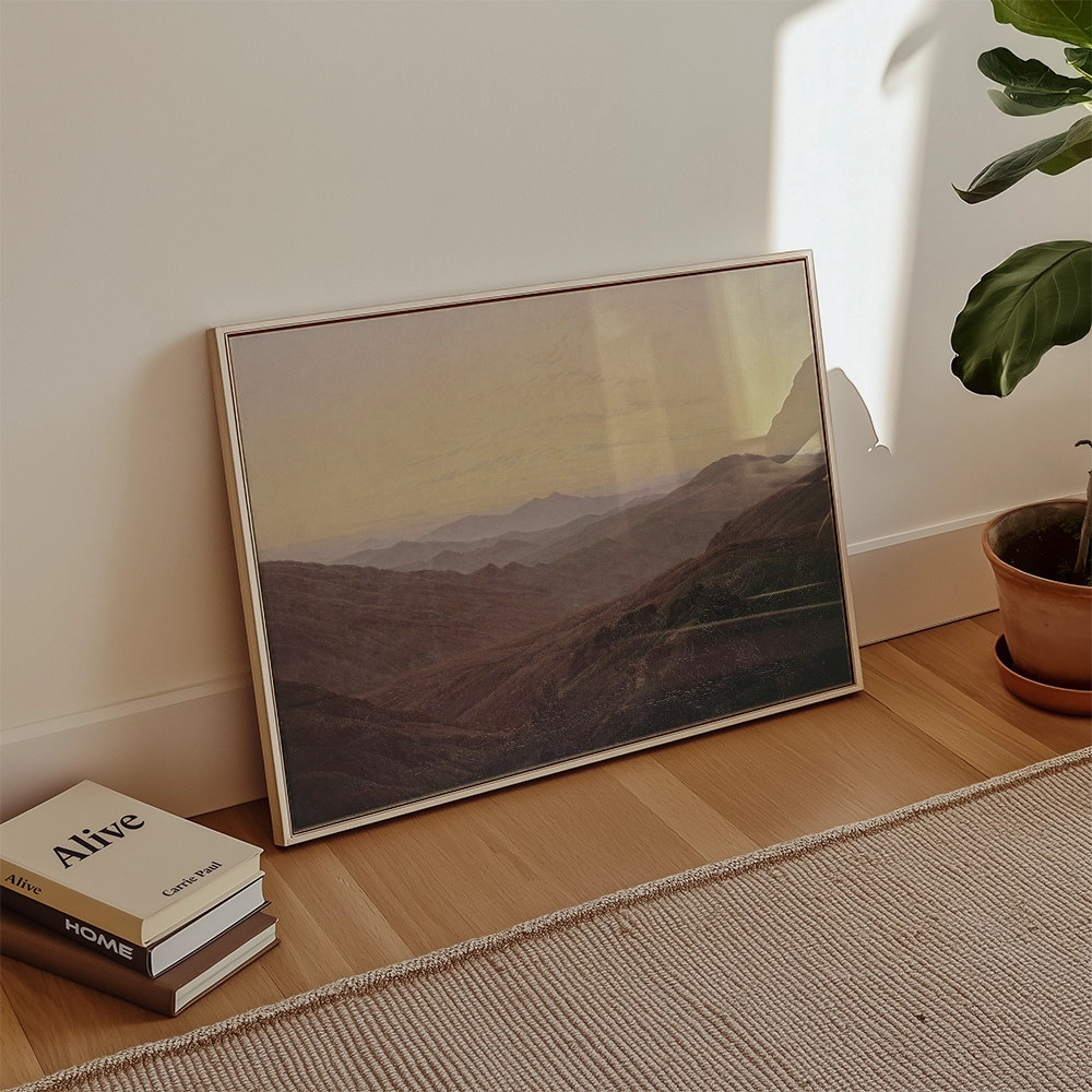 A framed landscape painting leaning against a wall in a modern interior with a plant and stacked books nearby.