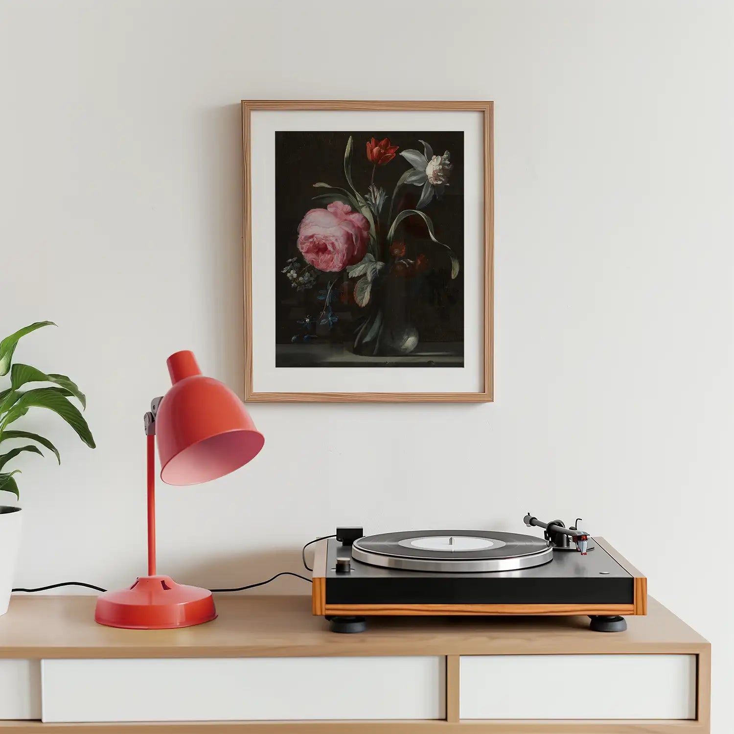 Floral painting in a wooden frame above a record player and a red lamp on a wooden table with a green plant in a white pot.