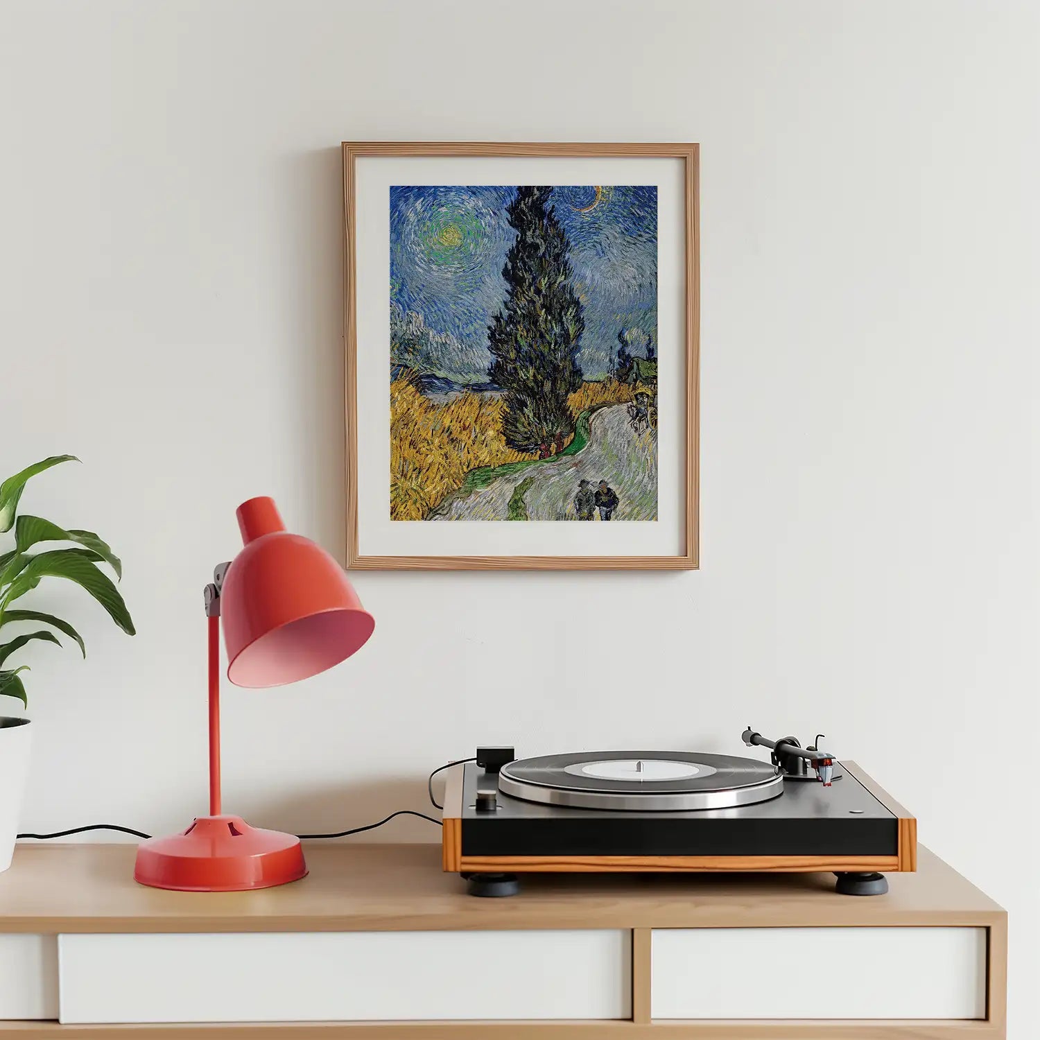 Framed painting of a cypress tree under a night sky, displayed above a vinyl record player and a red desk lamp, with a green plant in a white pot.