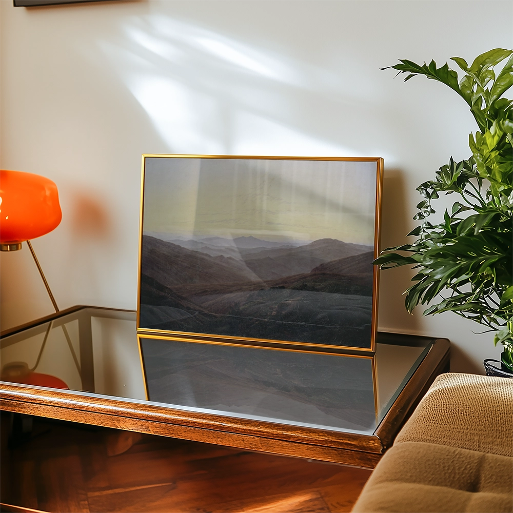 Framed landscape painting displaying rolling hills and mountains, positioned on a wooden table with a glass surface and a potted plant nearby.