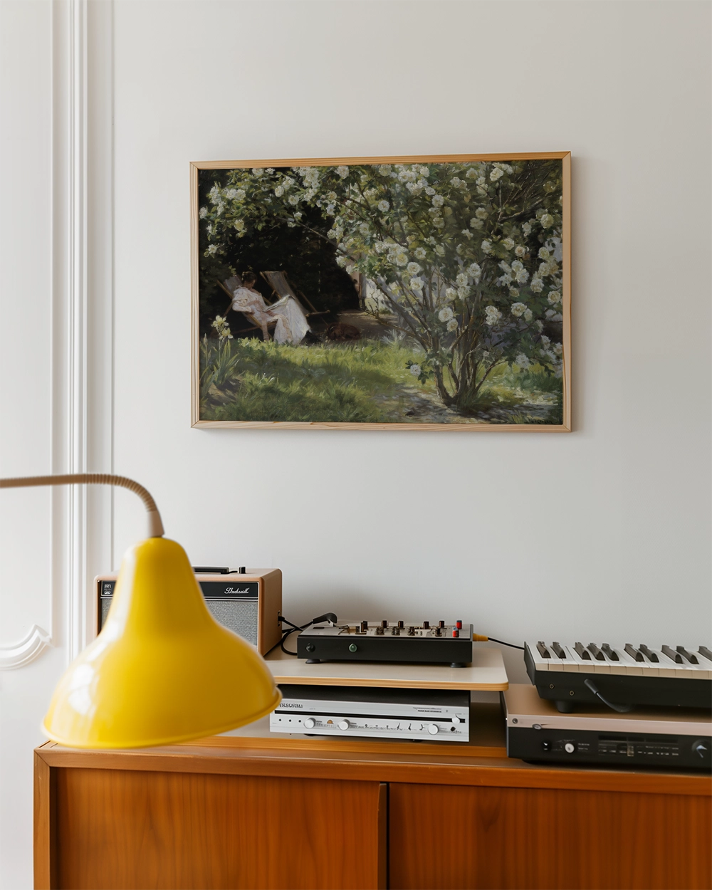 Painting of a woman reading under a flowering bush, framed and displayed on a white wall above a wooden cabinet with a yellow lamp and audio equipment.