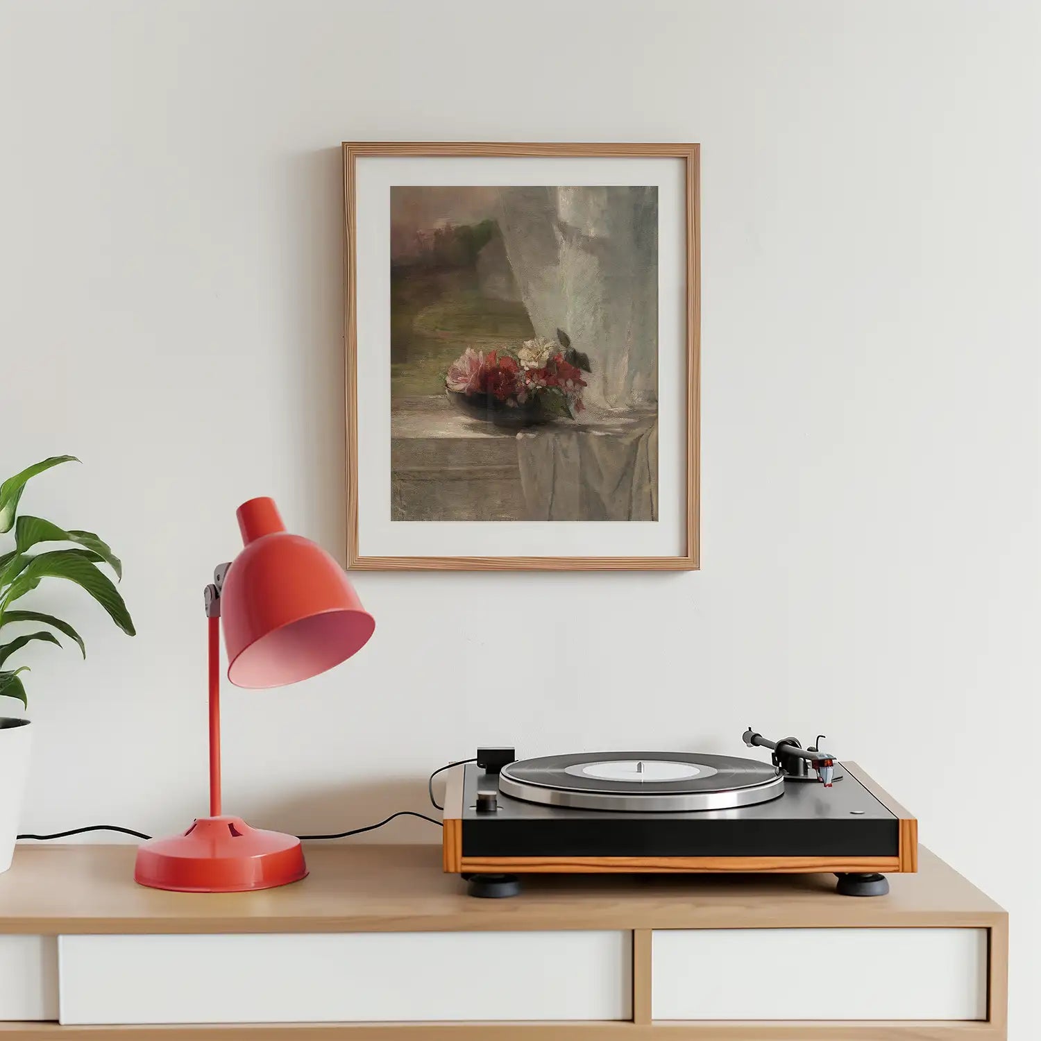 Painting in a wooden frame hanging on a wall above a record player and a red desk lamp on a wooden surface, with a green plant nearby.