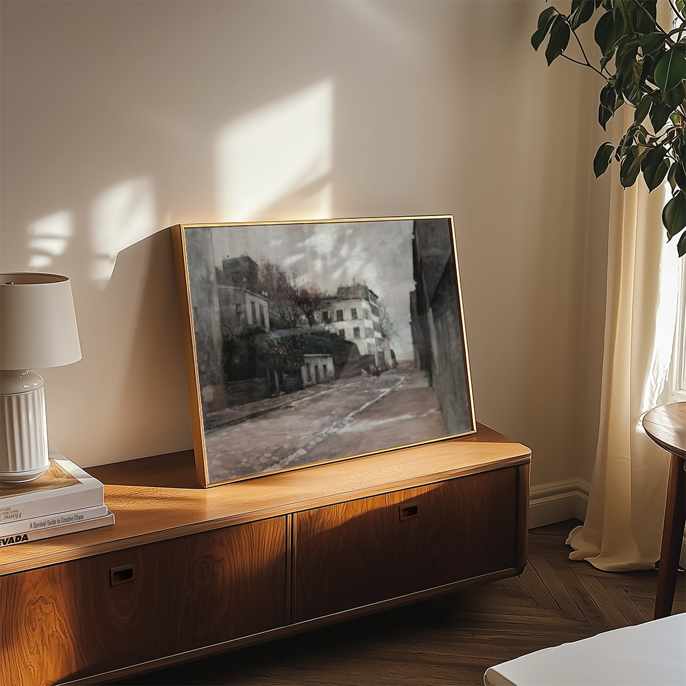 Painting of a serene street scene displayed in a gold frame on a wooden tabletop, with a lamp and plant nearby.