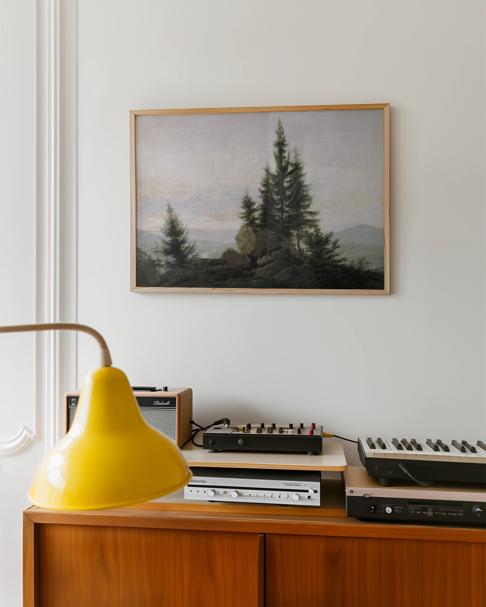 Framed painting of a serene landscape with evergreen trees and mountains, displayed above a wooden sideboard with a yellow lamp and audio equipment.