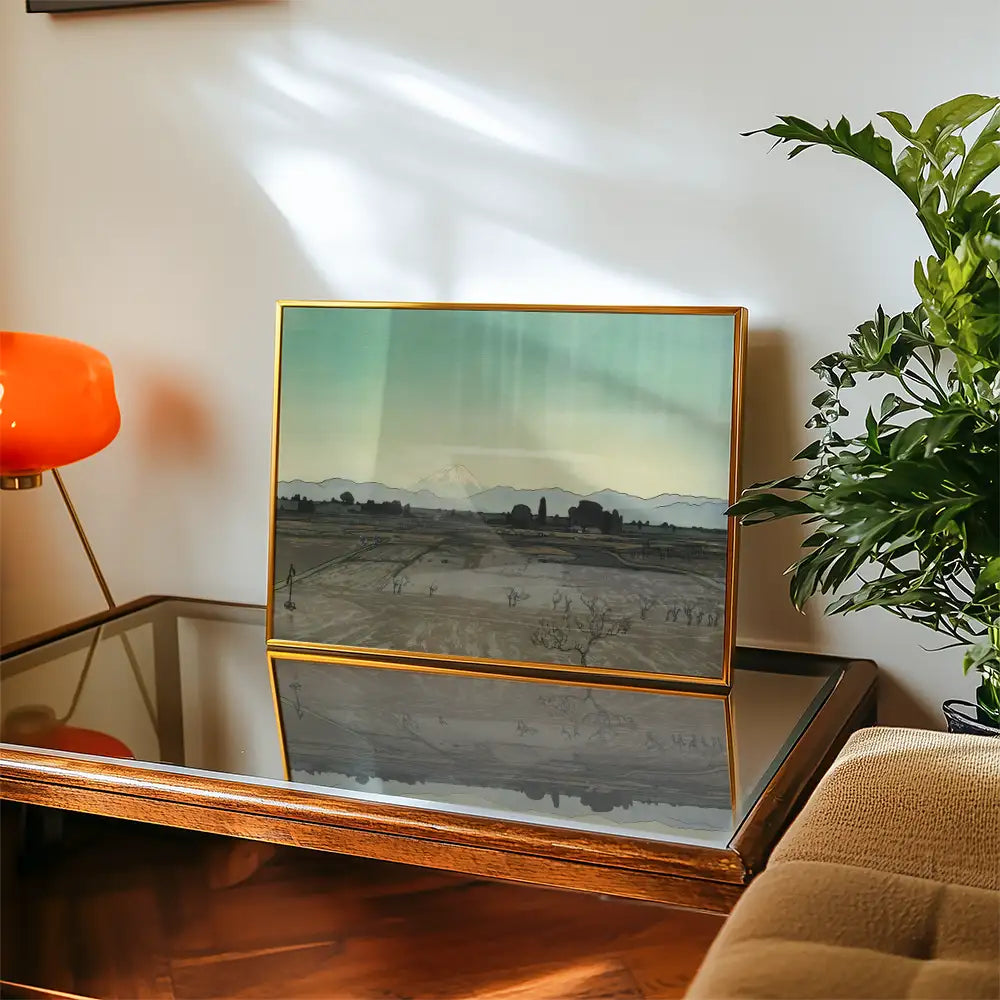 Framed landscape painting featuring mountains and a serene rural scene, displayed on a wooden table next to a modern orange lamp and greenery.