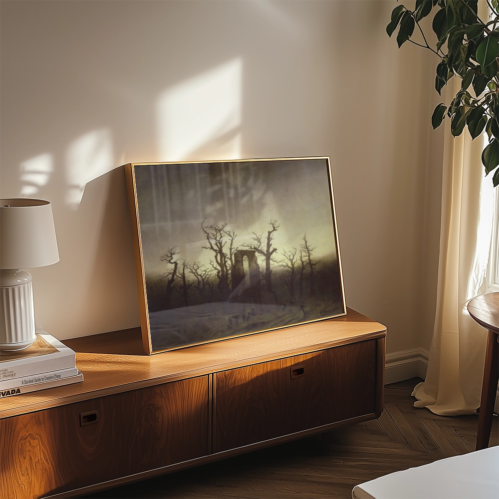 A framed painting of a moody landscape with bare trees, displayed on a wooden cabinet beside a lamp and decorative books in a softly lit room.