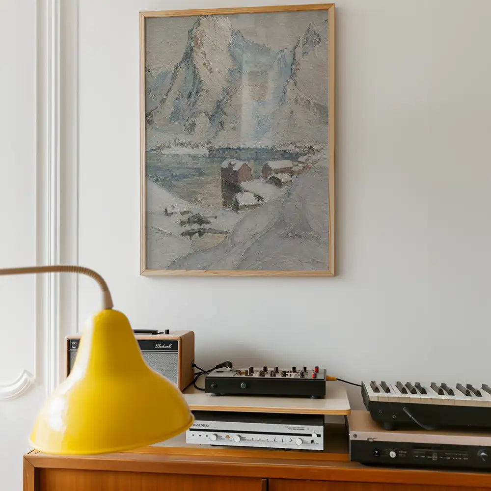 Framed painting of a snowy mountain landscape with a serene lake and small houses, displayed on a wall above a wooden side table with a yellow lamp and various audio equipment.