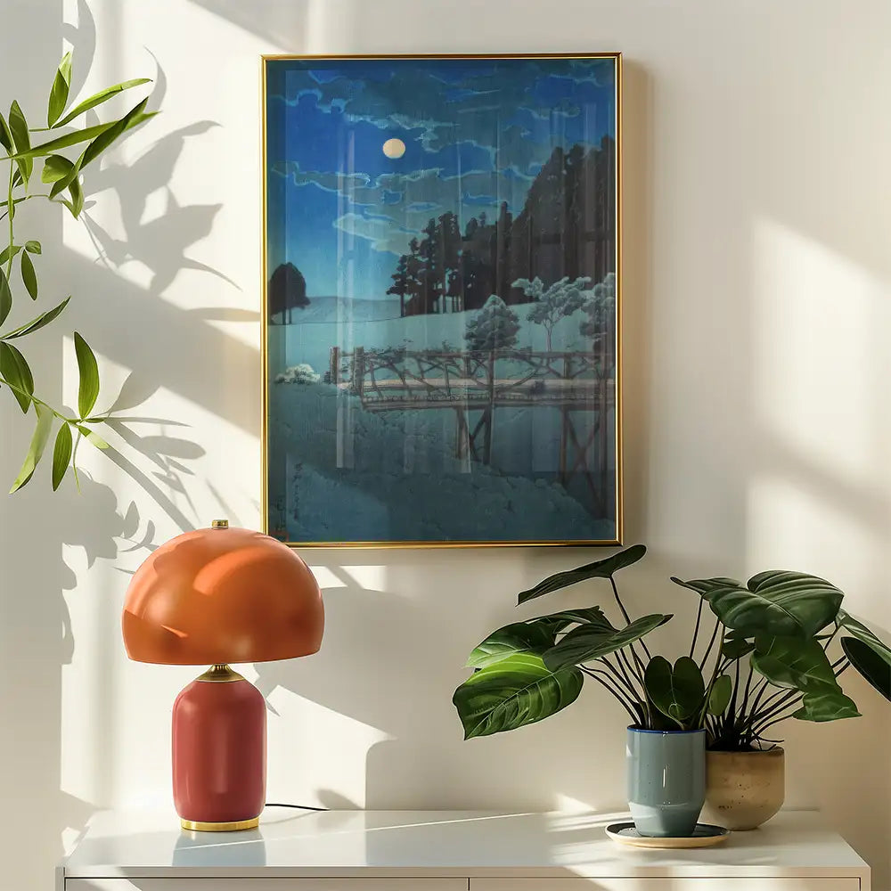 Artistic landscape painting in a gold frame, featuring a moonlit scene with trees and a bridge, displayed above a stylish orange lamp and potted plants on a modern white surface.