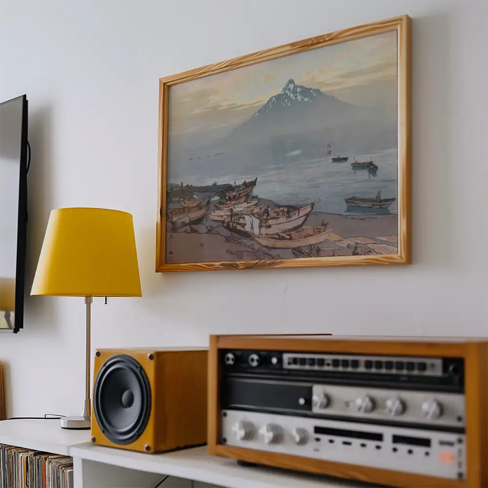 A framed painting of boats by the shore with a mountain backdrop, displayed above a yellow lamp and a vintage audio system.