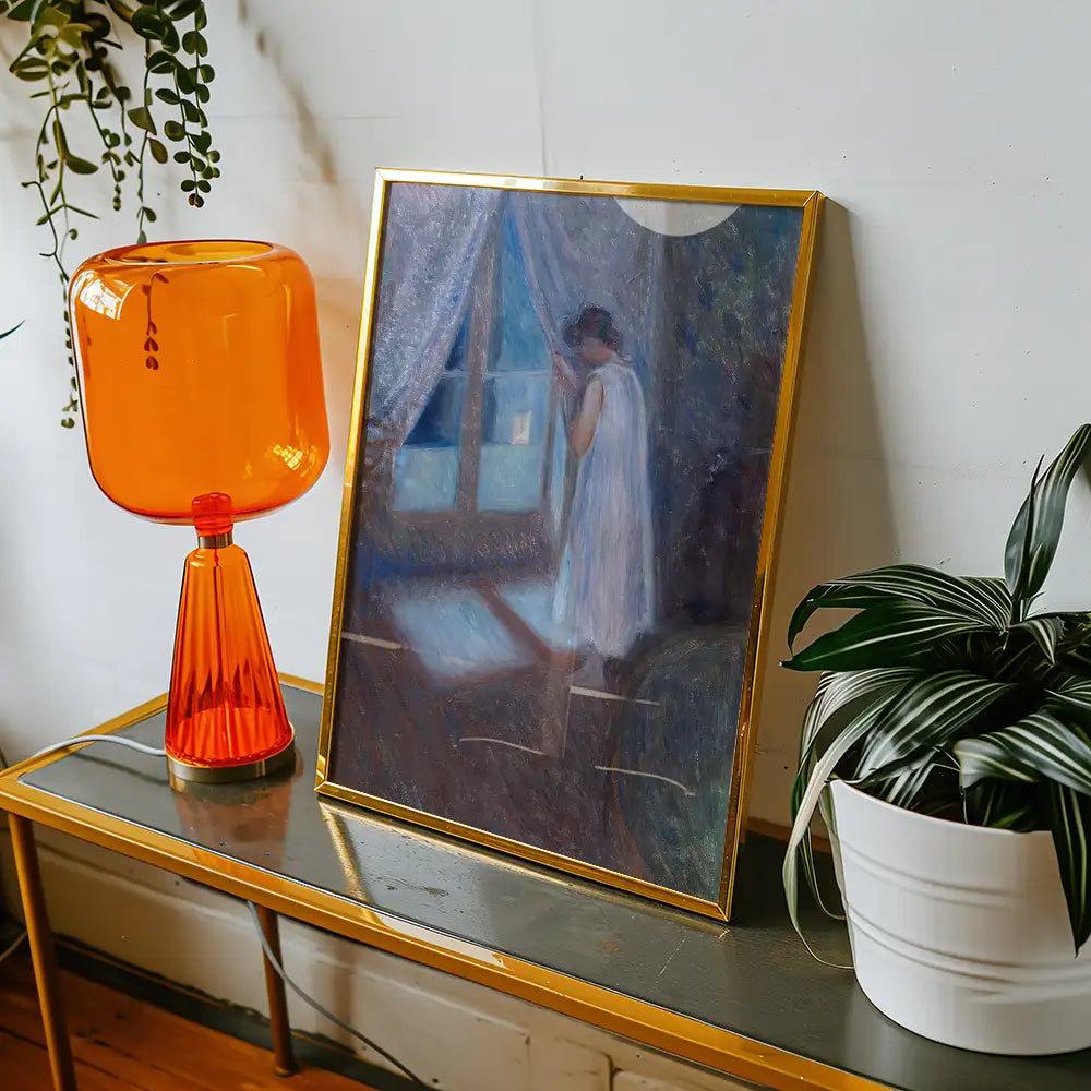 A framed painting of a woman in a white dress standing by a window, displayed on a table with an orange lamp and a potted plant.