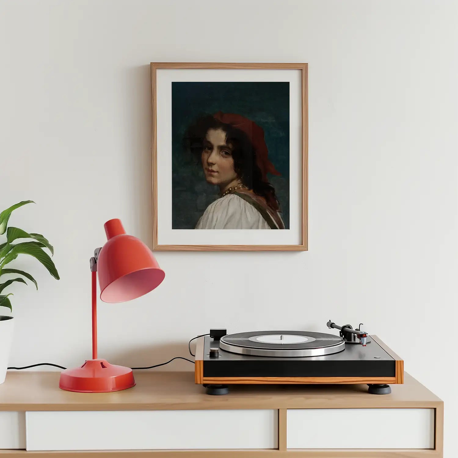 A framed portrait painting of a woman with a red headscarf, displayed on a wall above a record player and a pink desk lamp, with a green plant nearby.