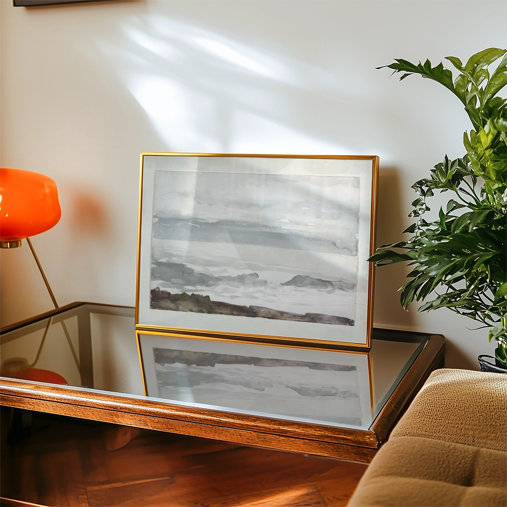 Framed watercolor painting of a seascape displayed on a glass table, with a decorative orange lamp and a green plant in the background.