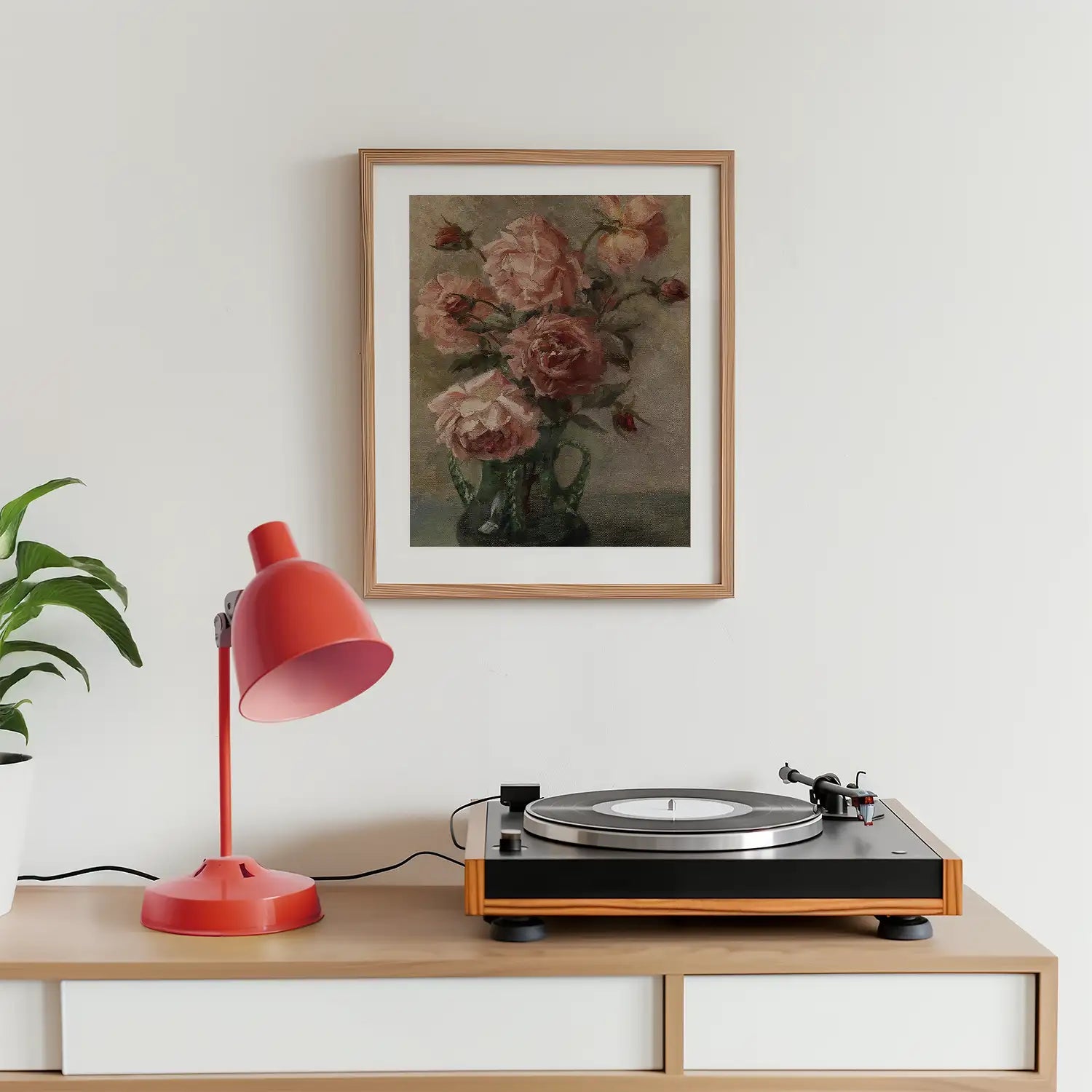 Framed painting of pink roses in a vase hanging above a wooden table with a red lamp and a record player.