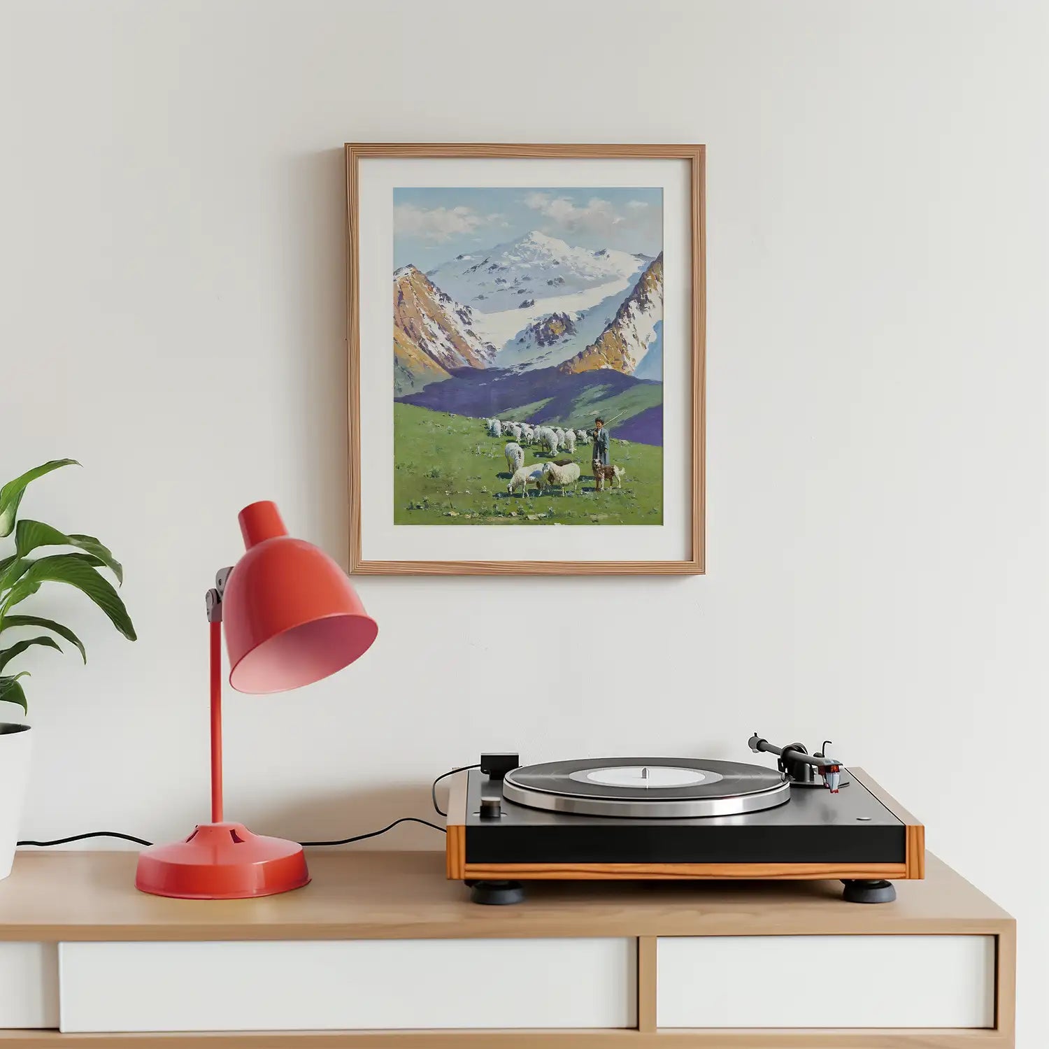 Framed painting of a pastoral landscape with mountains, sheep, and a farmer, displayed above a wooden table featuring a turntable and a red lamp.