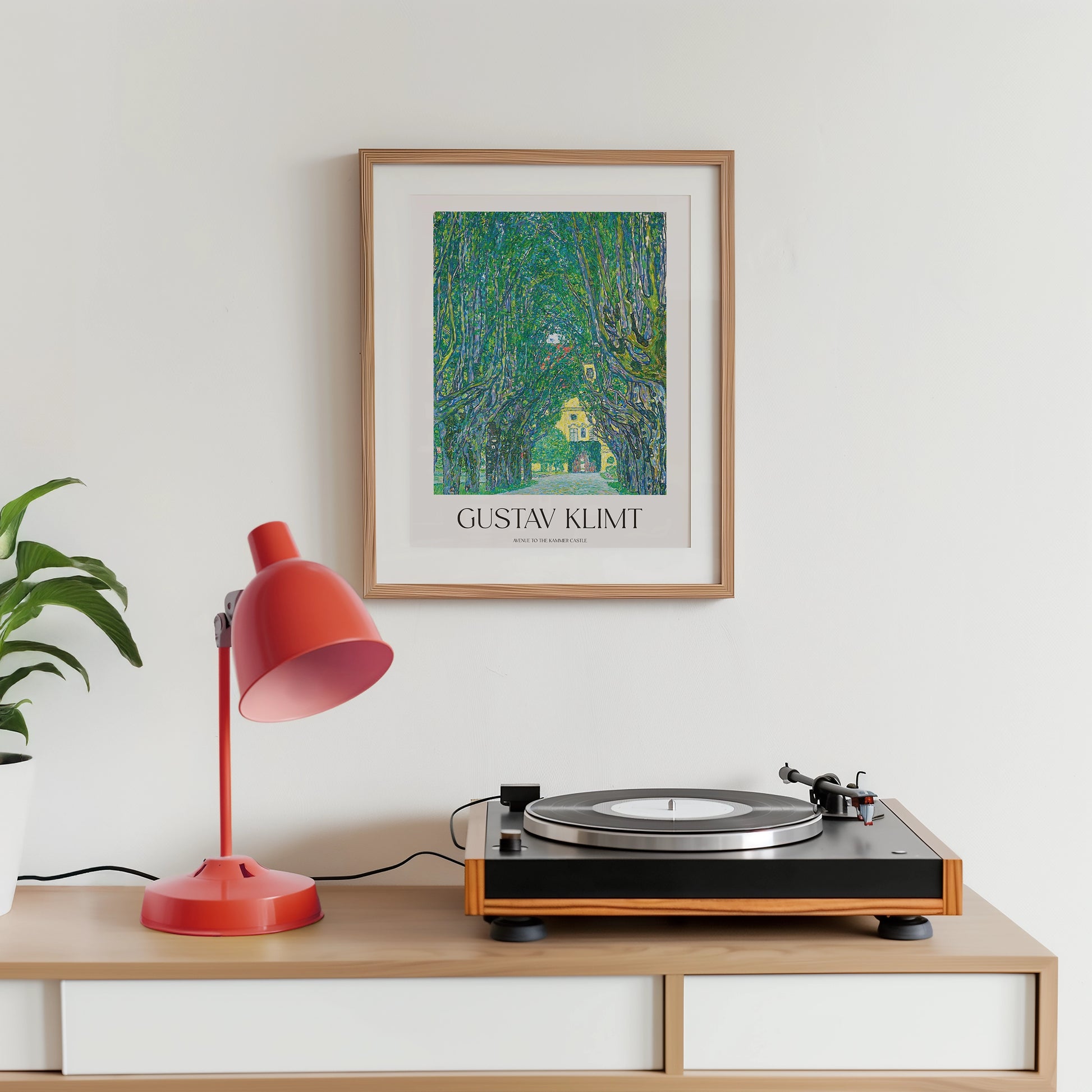Gustav Klimt painting poster displayed in a wooden frame on a wall above a record player with a red desk lamp and a plant on a storage unit.