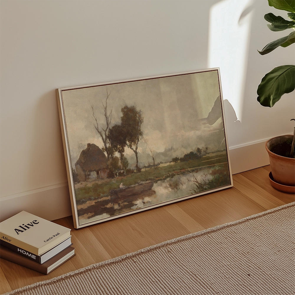 Framed painting of a serene landscape with trees and a cottage beside a river, leaning against a wall in a modern interior setting
