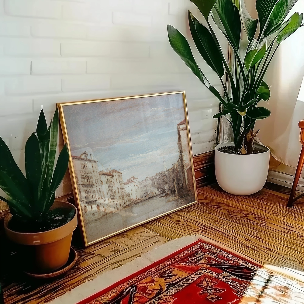 A framed painting of a serene canal scene leaning against a wall, accompanied by indoor plants and a decorative rug.