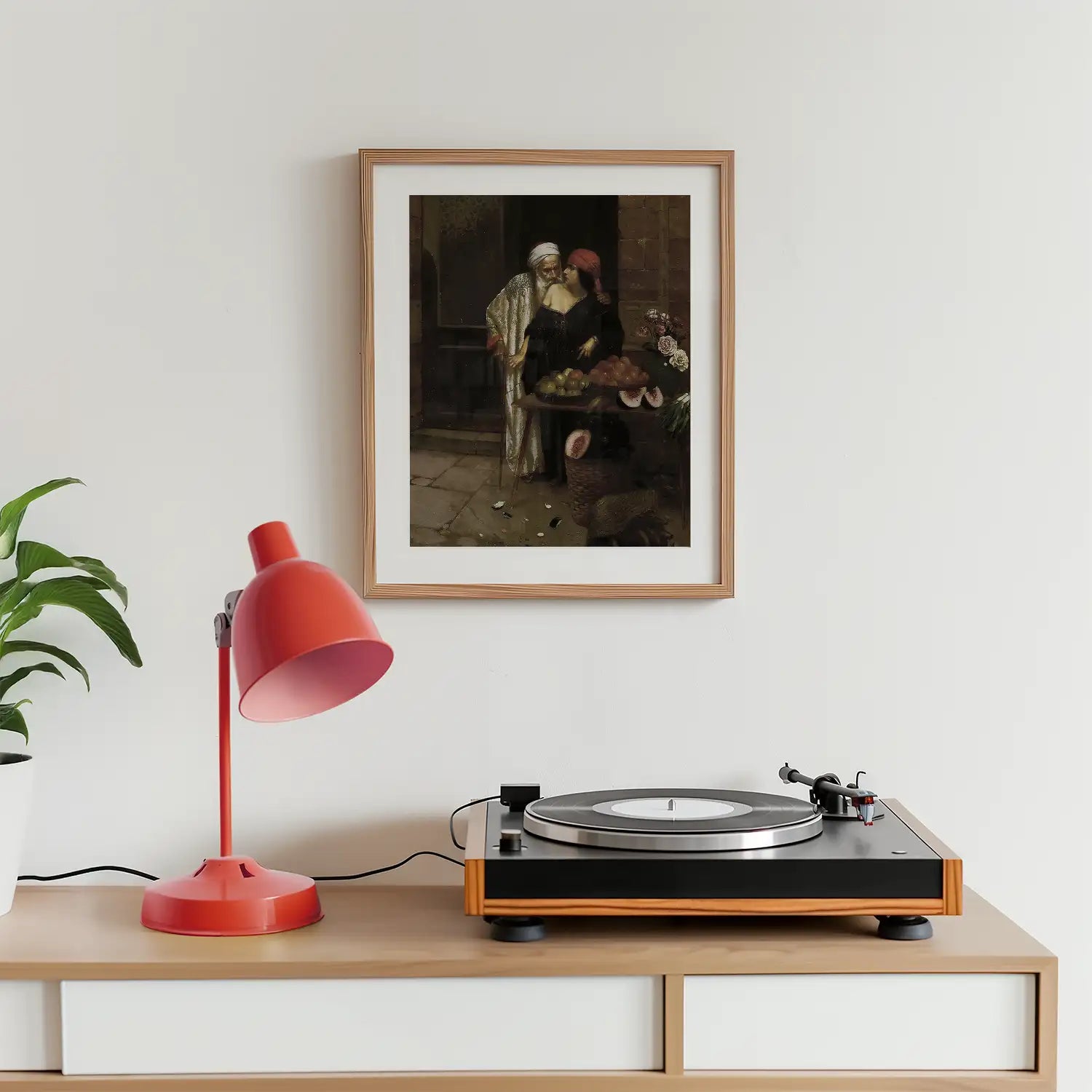 Framed painting of two figures in a market setting, displayed above a record player and a red desk lamp on a wooden surface, with a green plant in the background.
