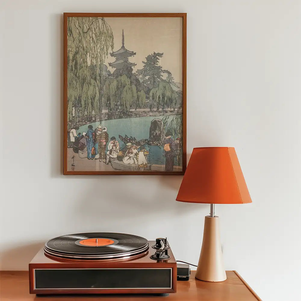 Framed vintage-style painting depicting a serene pond scene with people and trees, displayed above a record player and an orange lamp.