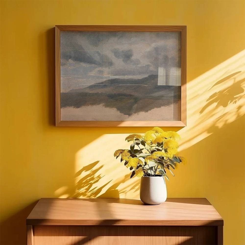 Framed landscape painting on a yellow wall above a wooden table with a vase of yellow flowers.