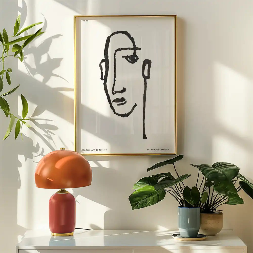 Modern art poster featuring abstract line drawing of a face, framed in gold, displayed in a bright interior with greenery and lamp.
