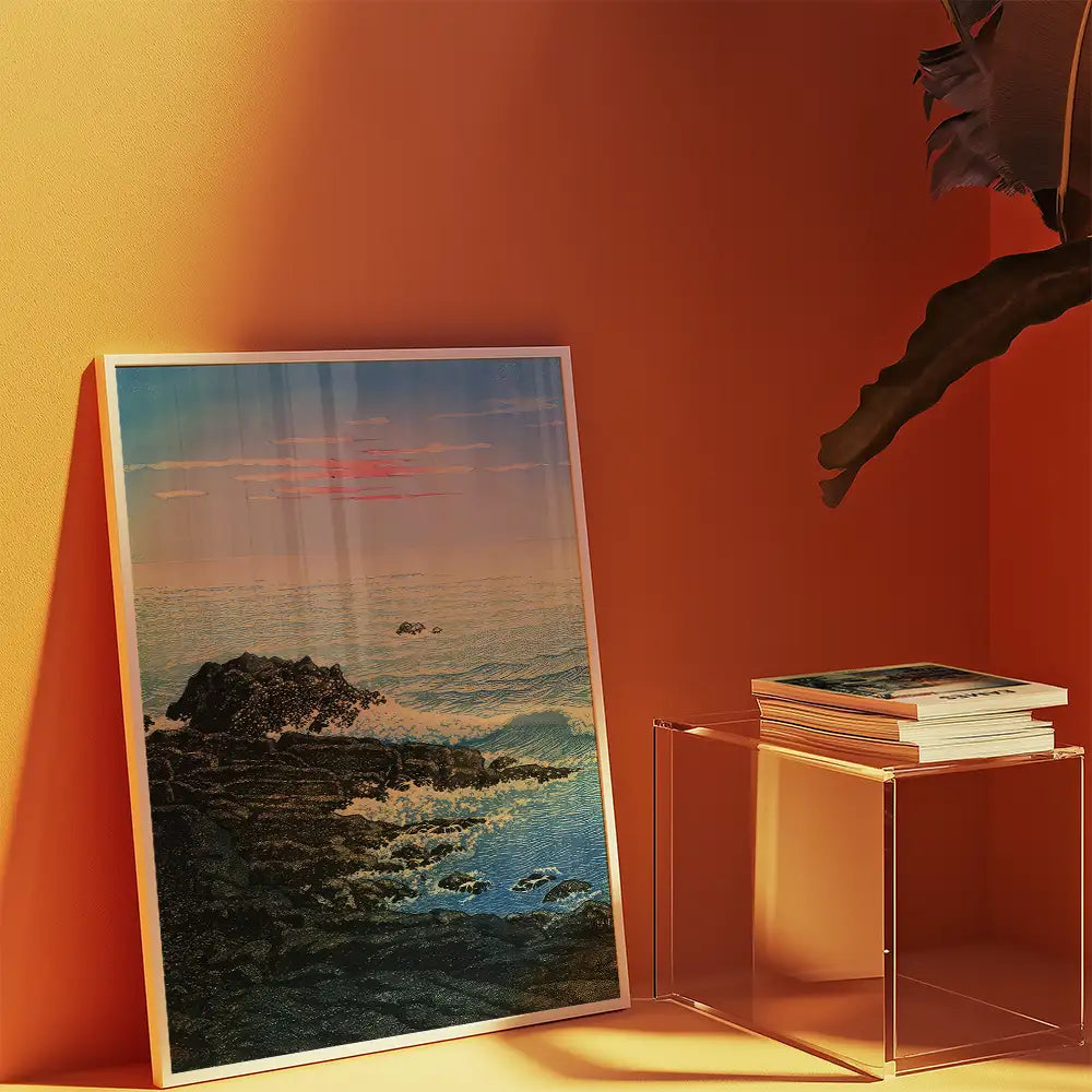 Framed coastal landscape painting with ocean waves and sunset, displayed on an orange wall next to a stack of art books on a clear acrylic table.