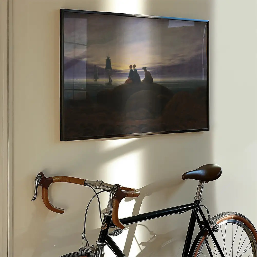 Framed painting depicting a romantic sunset scene with two figures sitting on rocks by the water, with a bicycle leaning against the wall below the artwork.