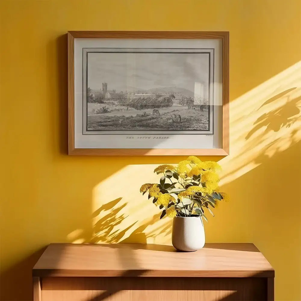 Framed black and white landscape painting titled "The South Parade" hanging on a yellow wall above a wooden cabinet with a vase of yellow flowers.