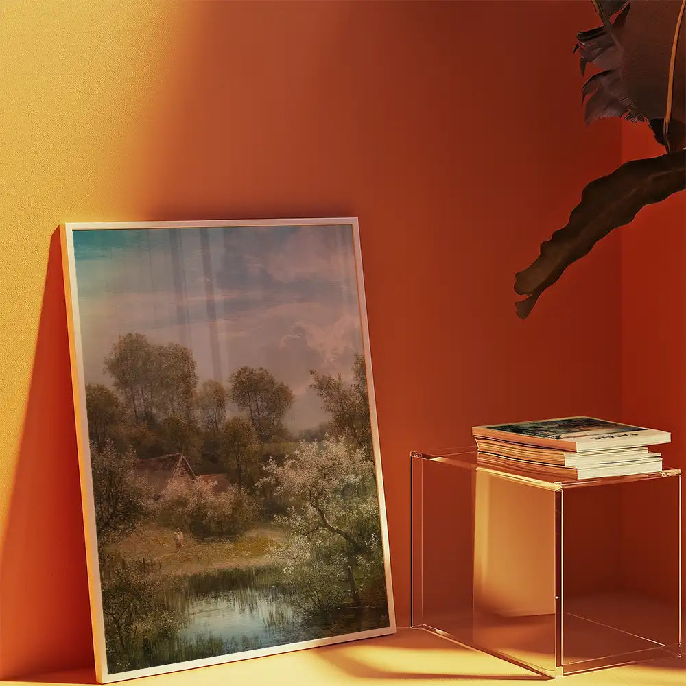 Framed landscape painting featuring trees and a serene body of water, displayed against an orange wall next to a stack of books on an acrylic table.