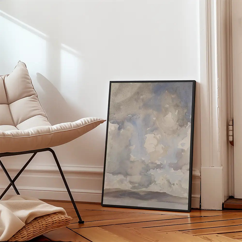 Watercolor abstract painting in a black frame leaning against a white wall next to a modern chair and a blanket, with wooden flooring.