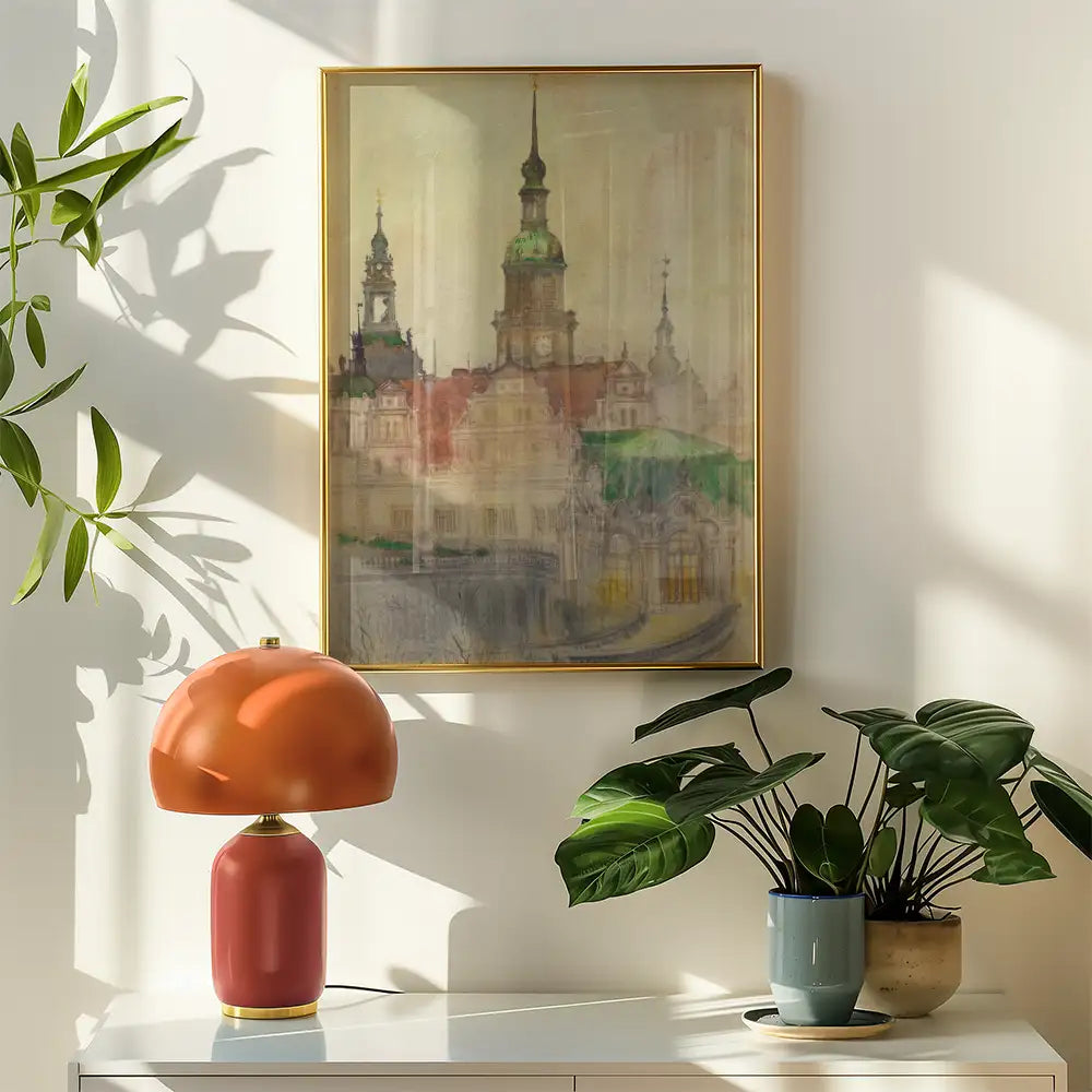Painting of a historic building in soft colors, framed in gold, displayed on a wall next to a stylish orange lamp and potted plants.