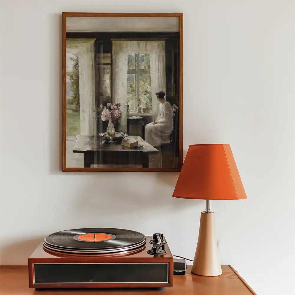 Painting of a woman sitting by a window with a vase of flowers, displayed above a record player and an orange lamp on a wooden table.