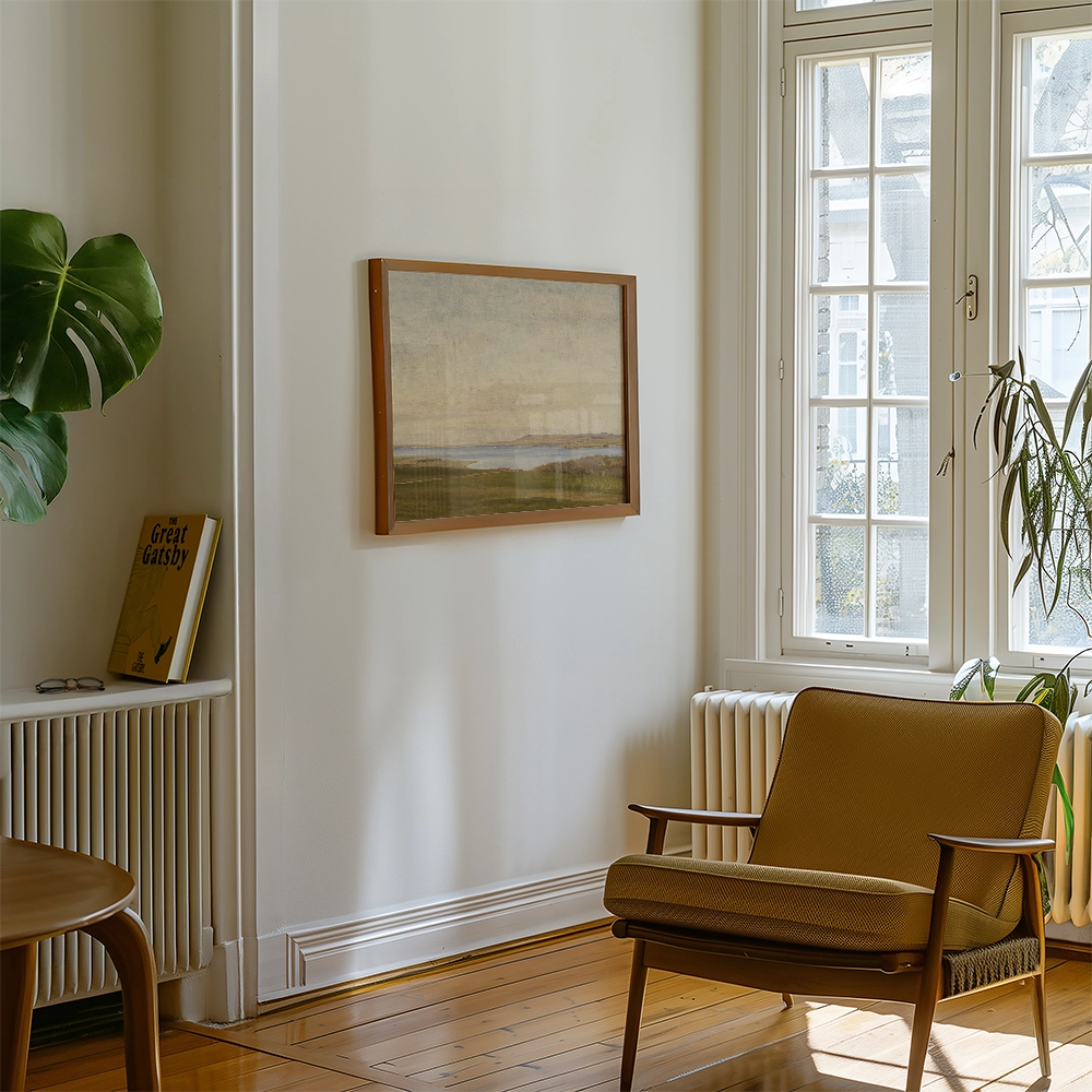 A landscape painting in a wooden frame hanging on a wall in a cozy room with a brown armchair and a plant.