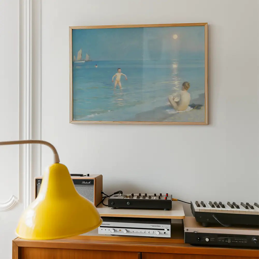 Framed painting of two figures in shallow water by the beach with a serene sky and distant sailboats, displayed above a wooden sideboard with a yellow lamp and vintage audio equipment.