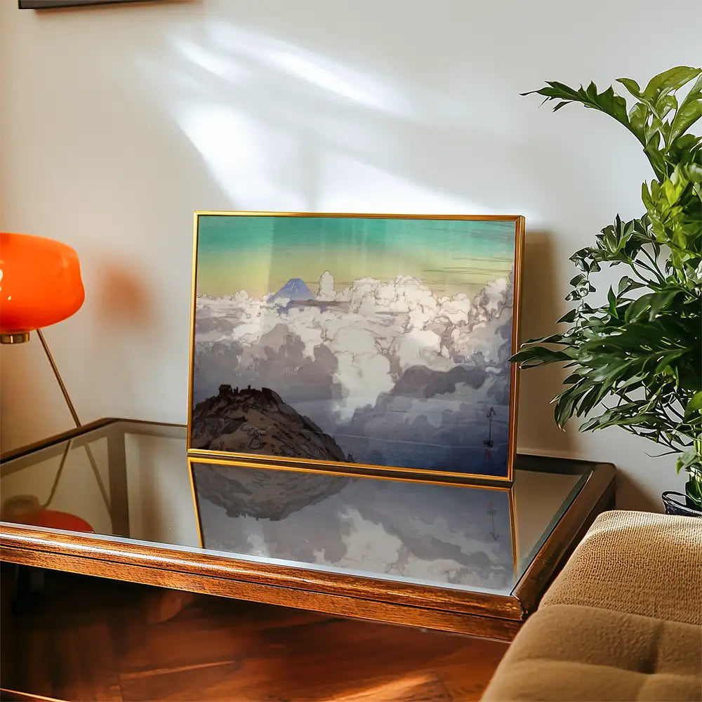 Framed painting of clouds and mountains displayed on a coffee table with a modern lamp and plant in the background.