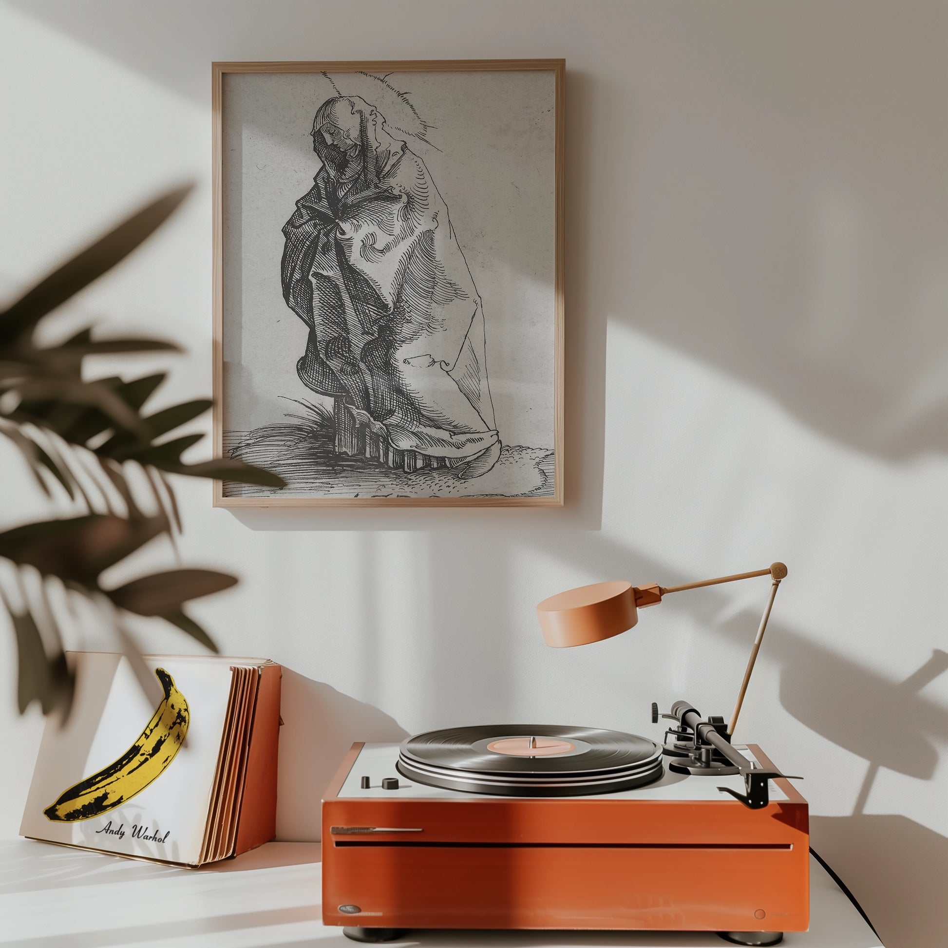 Framed black and white artwork of a cloaked figure on a wall above a stylish orange record player with a lamp and a banana-themed record cover nearby.