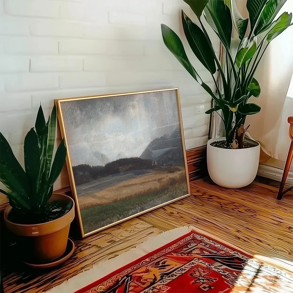 A framed landscape painting resting on a wooden floor next to a potted plant in a bright, cozy room.