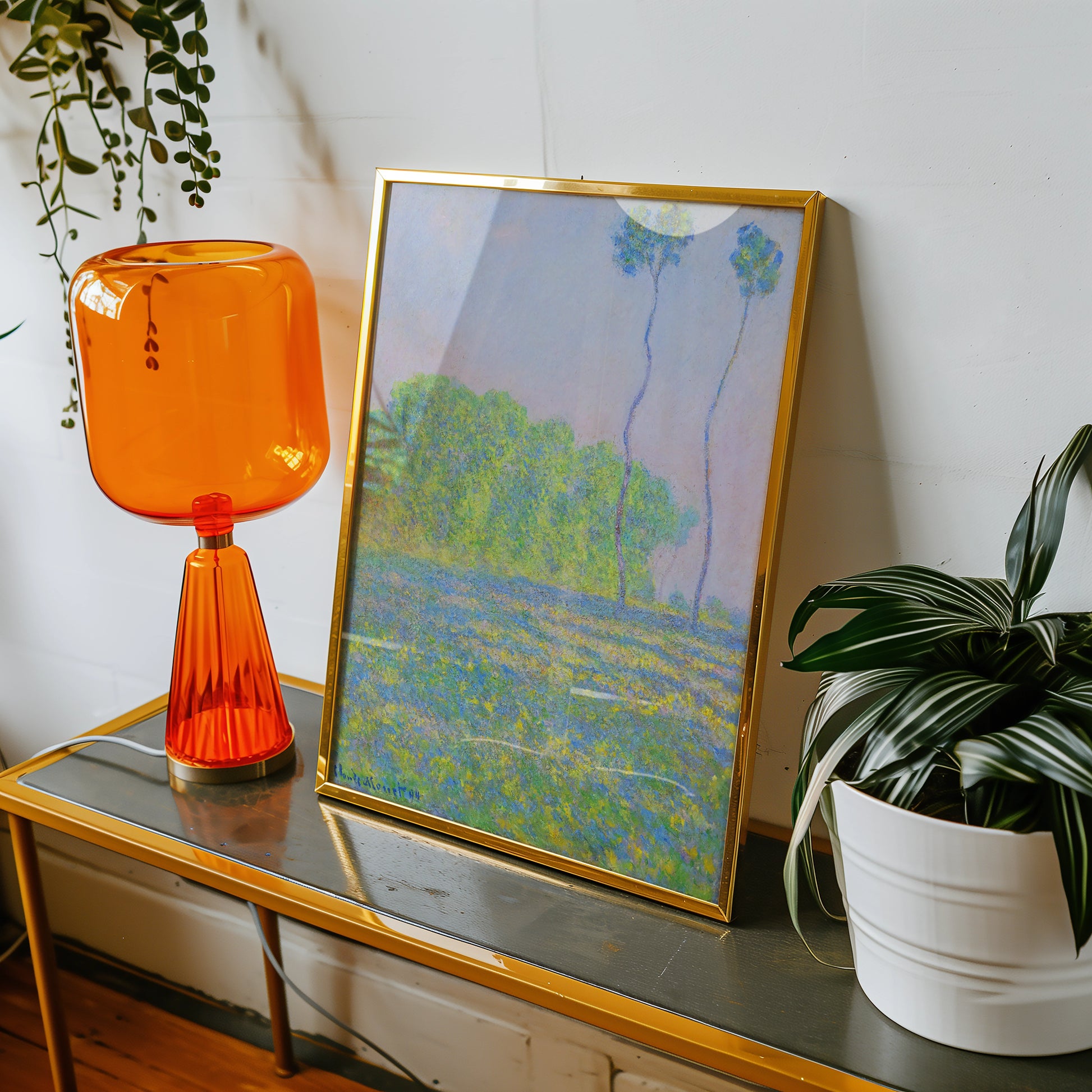 Framed painting featuring a colorful landscape with trees, displayed on a table alongside an orange lamp and a potted plant.