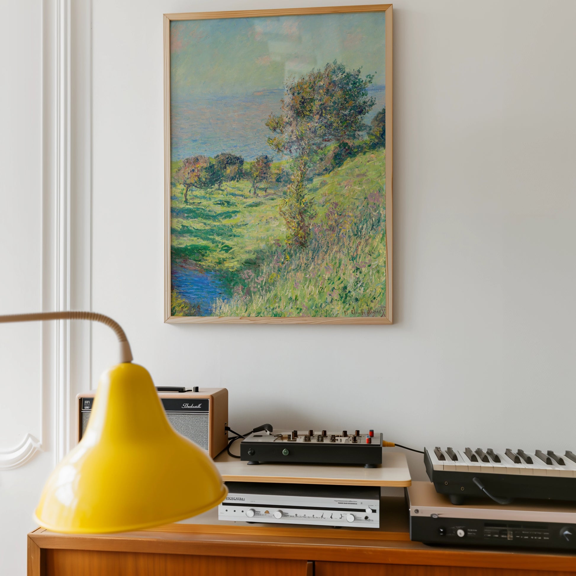 Painting of a serene landscape featuring a grassy hill and trees by the ocean, framed and displayed on a wall above a wooden console with audio equipment and a yellow lamp.