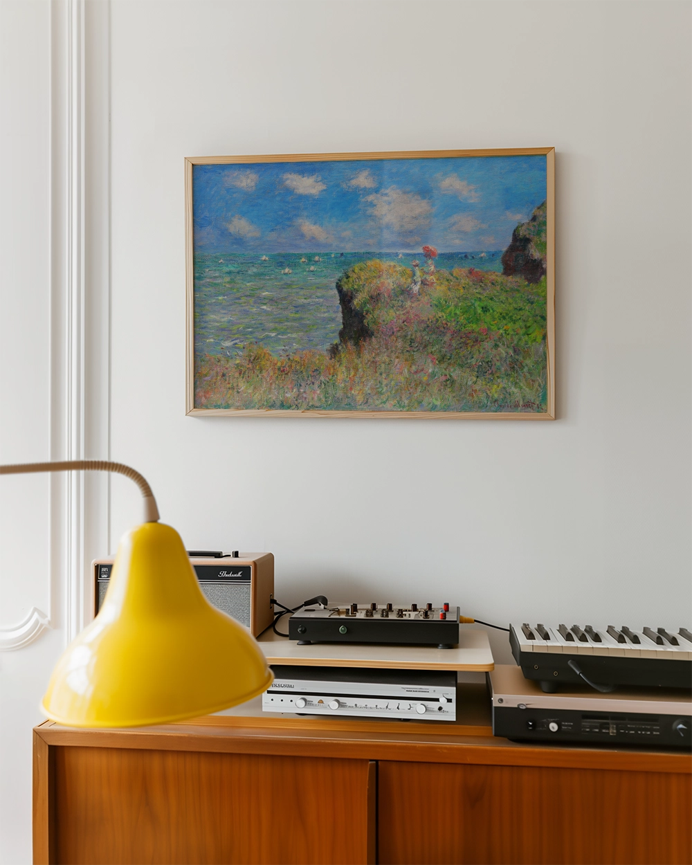 Painting of a seaside landscape with boats on the water, framed in gold, displayed on a wall above a wooden console with a yellow lamp and audio equipment.